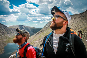 Two Caucasian men, wearing caps, with brown beards, backpacks, glasses, and Nöz sunscreen.